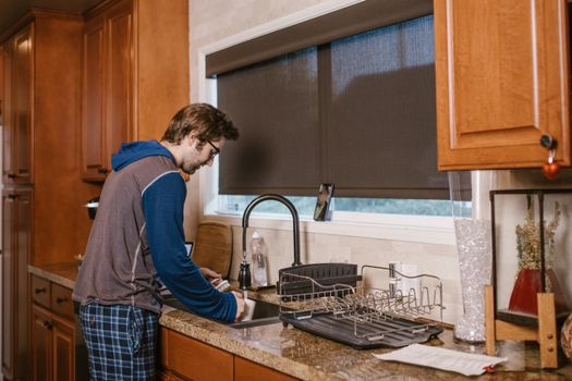 A man washing dishes at a kitchen sink with wooden cabinets and modern appliances.