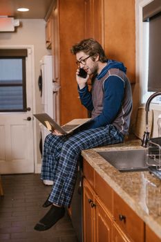 Caucasian man in pajamas working remotely on laptop while talking on phone in kitchen.