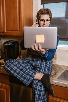 A man in pajamas using a laptop and phone in the kitchen, illustrating remote work.