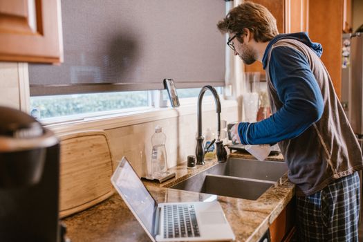 A man multitasks by washing dishes and working remotely at a kitchen sink with a laptop.