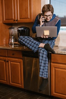A man in pajamas multitasking on a laptop and phone in the kitchen, showcasing a lifestyle moment.