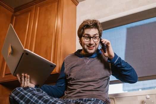 Caucasian man using laptop and phone for remote work at home, relaxed and multitasking.