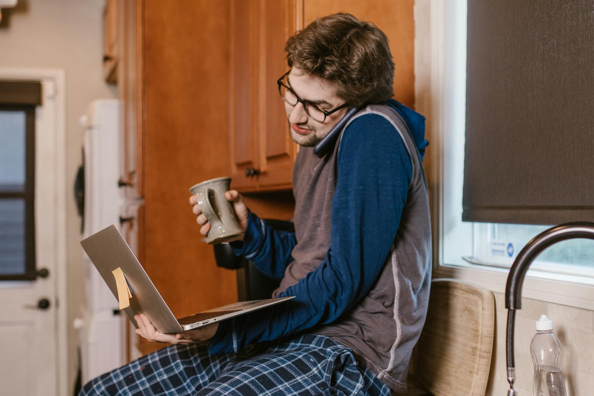 Caucasian man multitasking in kitchen, balancing phone call and laptop work with a mug in hand.