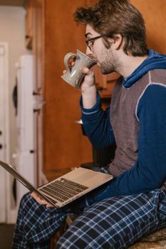 Caucasian man in loungewear drinking coffee while using a laptop for online learning.