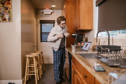 Caucasian man buttoning shirt in modern kitchen near laptop and coffee maker.