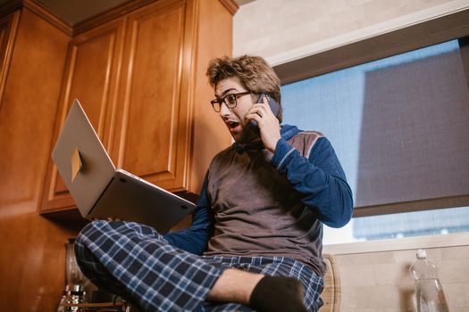 A surprised man in pajamas multitasking with a laptop and phone indoors.