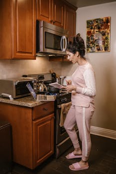 A woman in a kitchen cooking while referencing a laptop and notebook.