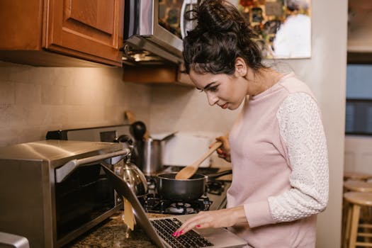 Young woman multitasking in kitchen, cooking while using laptop. Ideal for remote work or distance learning themes.