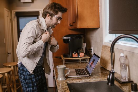 A man in home kitchen buttoning shirt while attending an online meeting on laptop.