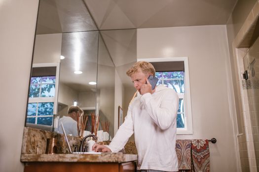 Adult man multitasking by brushing teeth and talking on phone in modern bathroom.