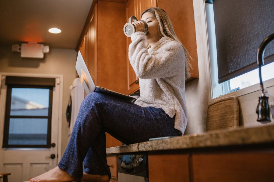 Casual morning scene of a woman enjoying coffee and using a laptop sitting on a kitchen counter.