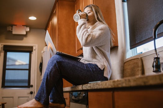 Casual morning scene of a woman enjoying coffee and using a laptop sitting on a kitchen counter.