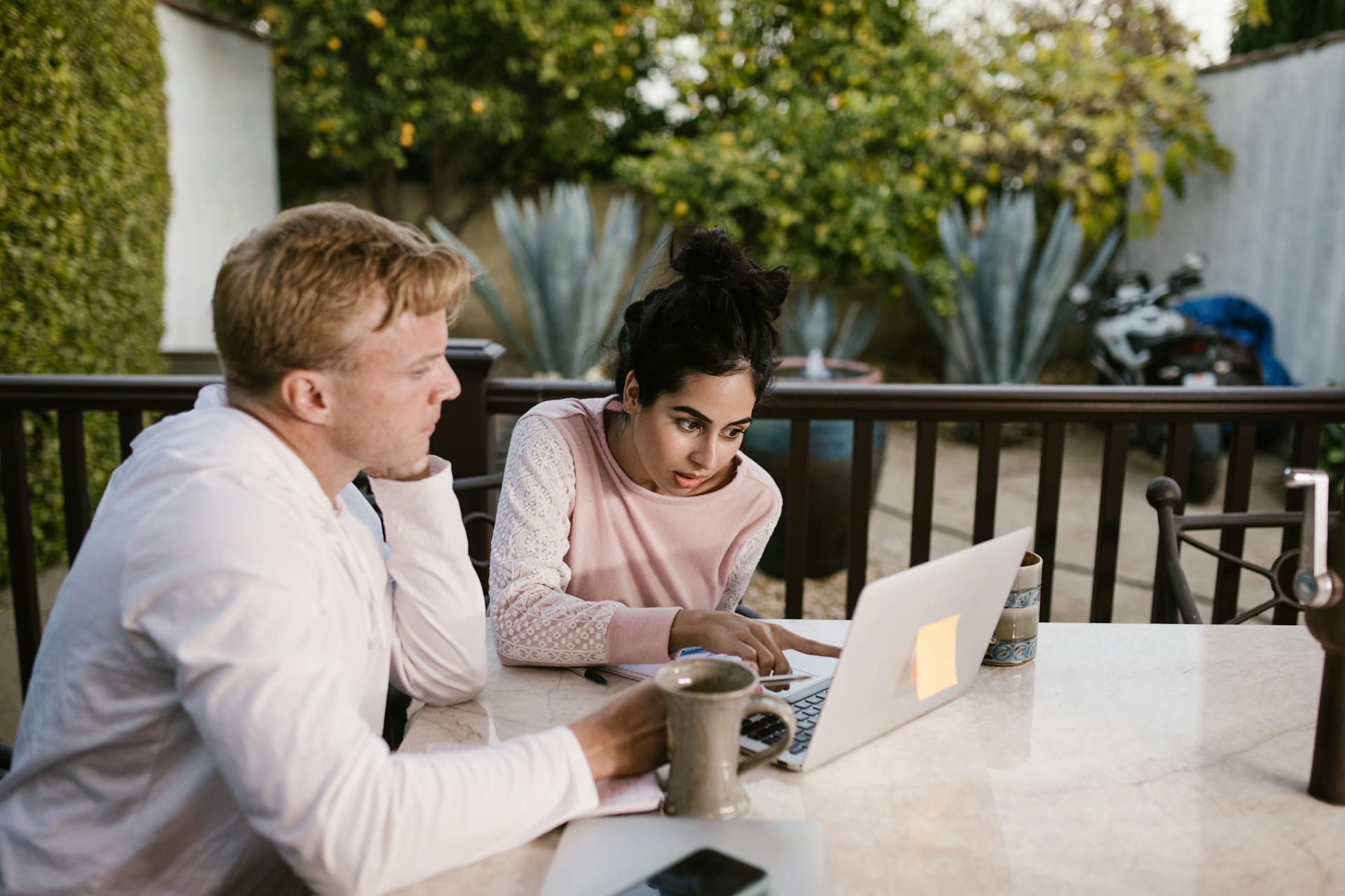 Two adults collaborating on a laptop, seated outdoors with coffee on the table.