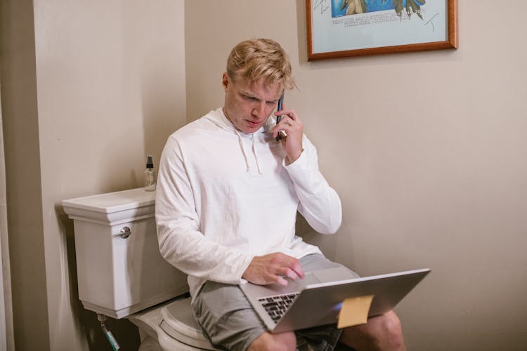 Man Sitting On Toilet Using Laptop And Phone