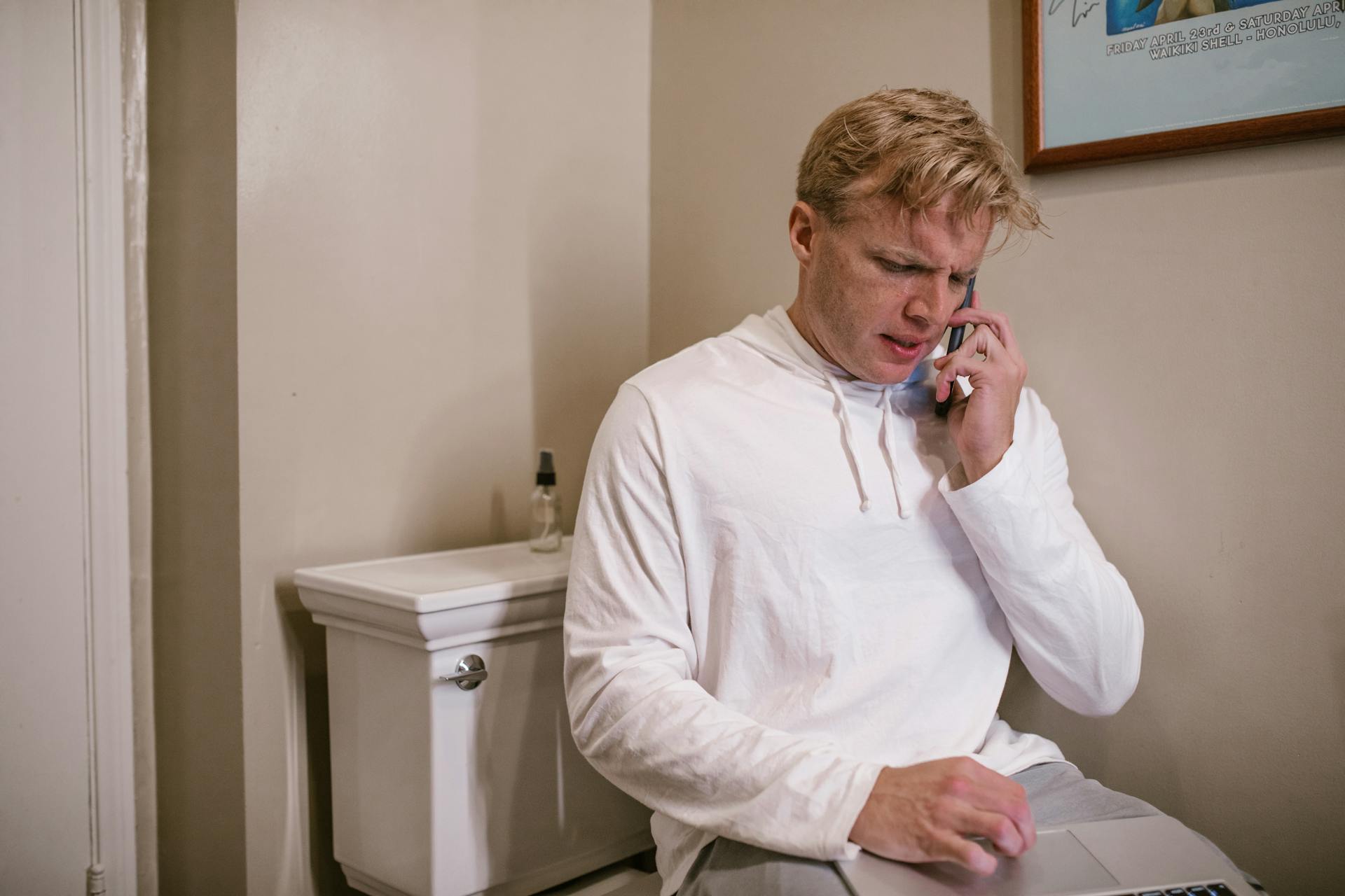 Man multitasking on phone and laptop in bathroom, showcasing modern life.