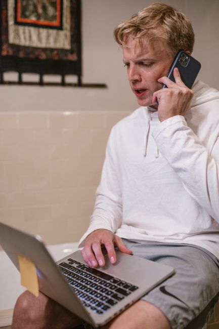 Adult man multitasks with laptop and phone wearing a white hoodie indoors. Adult man multitasks with laptop and phone wearing a white hoodie indoors.