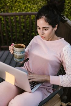 Woman in casual attire using a laptop while enjoying a cup of coffee outdoors.