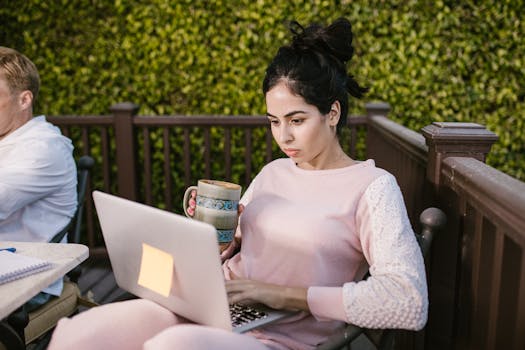 Woman focused on laptop work outdoors, holding a coffee mug. Enjoying remote work in cozy setting.