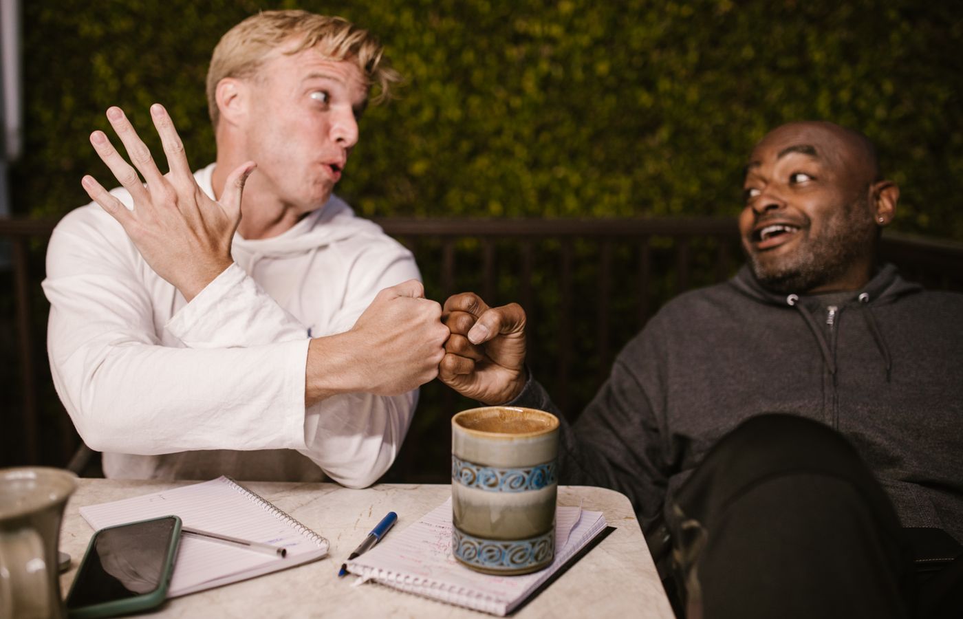 Two friends share a joyful fist bump while enjoying coffee outdoors, highlighting friendship.