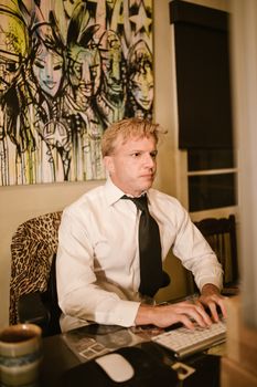 A man in formal attire works diligently at his computer under vibrant graffiti artwork inside his office.