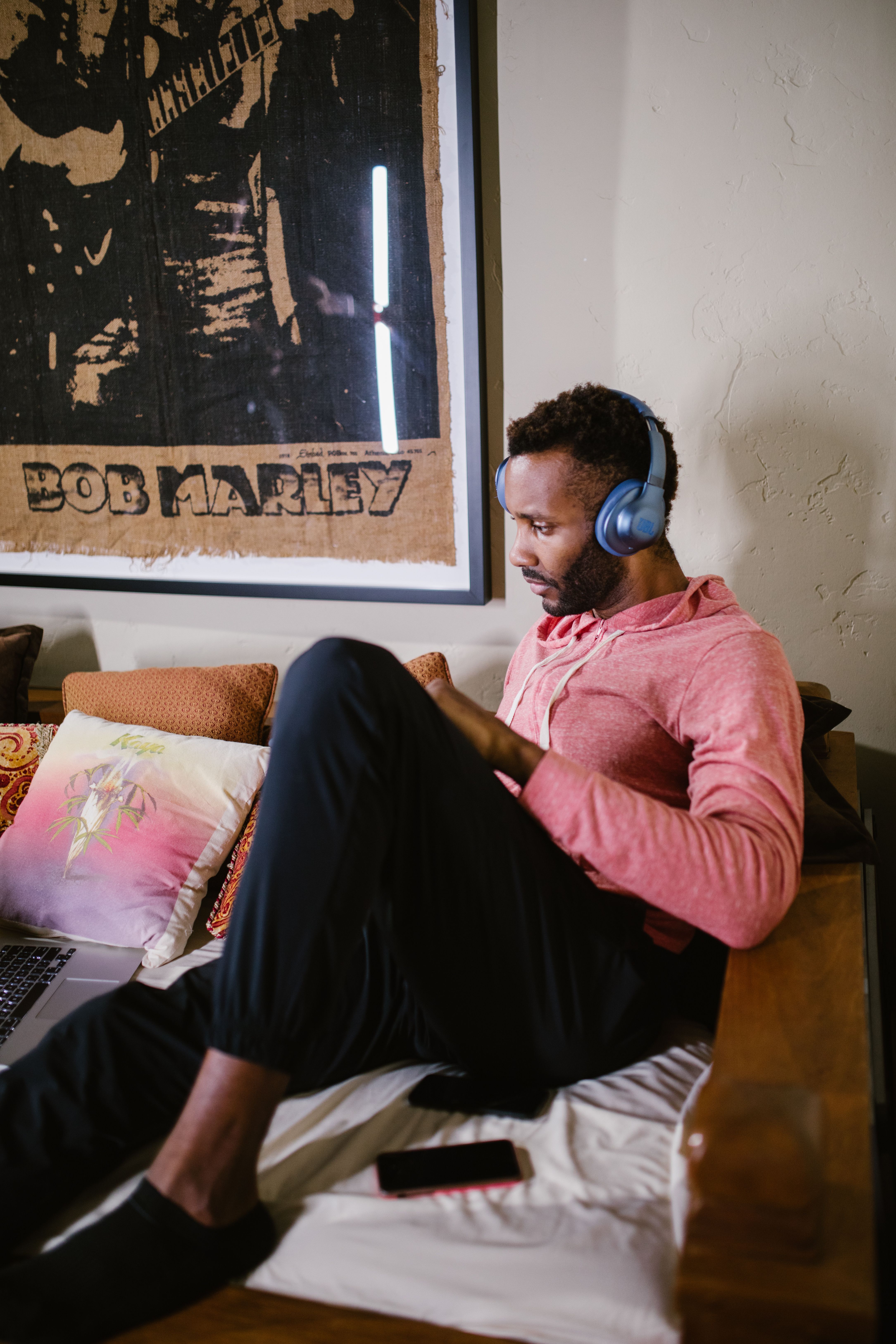 A man in a pink hoodie uses a laptop with headphones in a cozy setting featuring a Bob Marley poster.
