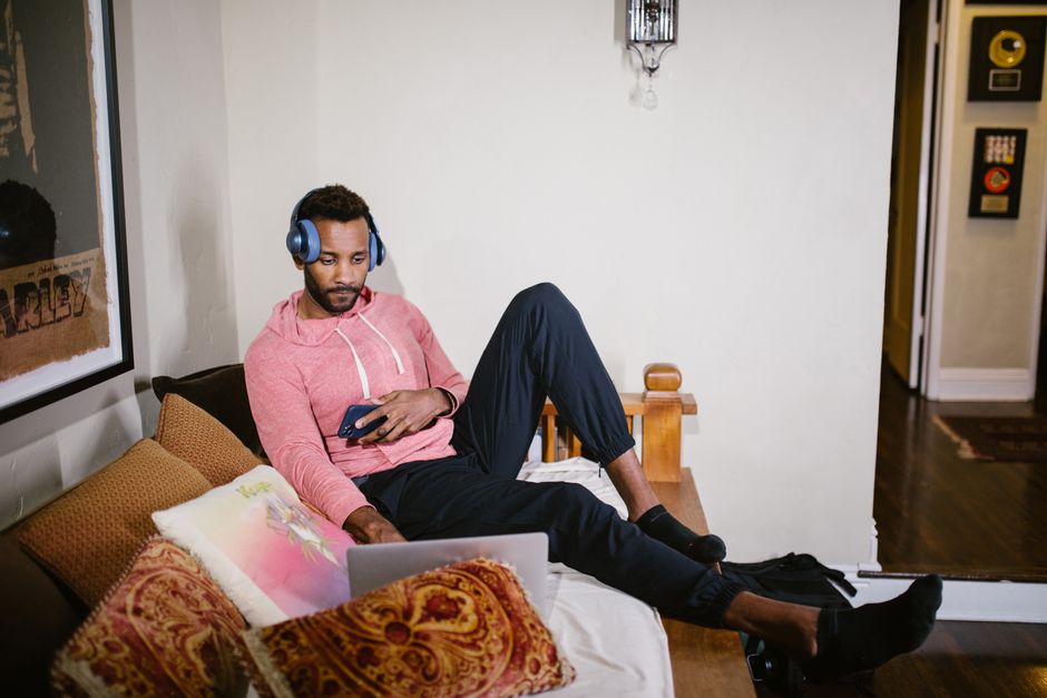 A man wearing headphones relaxes with laptop and phone on a cozy couch indoors.