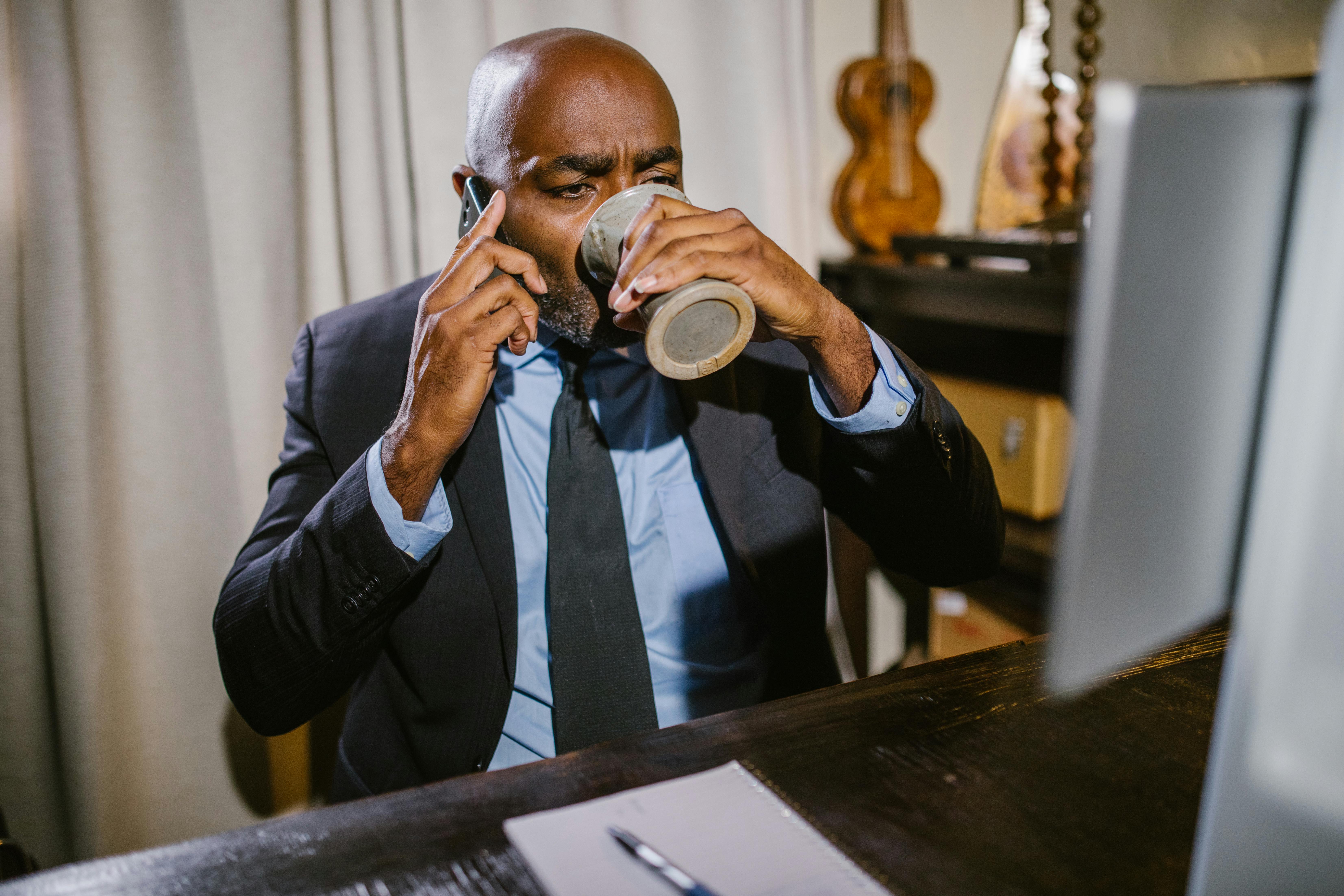 A man in a suit sips coffee and talks on the phone while working at home.