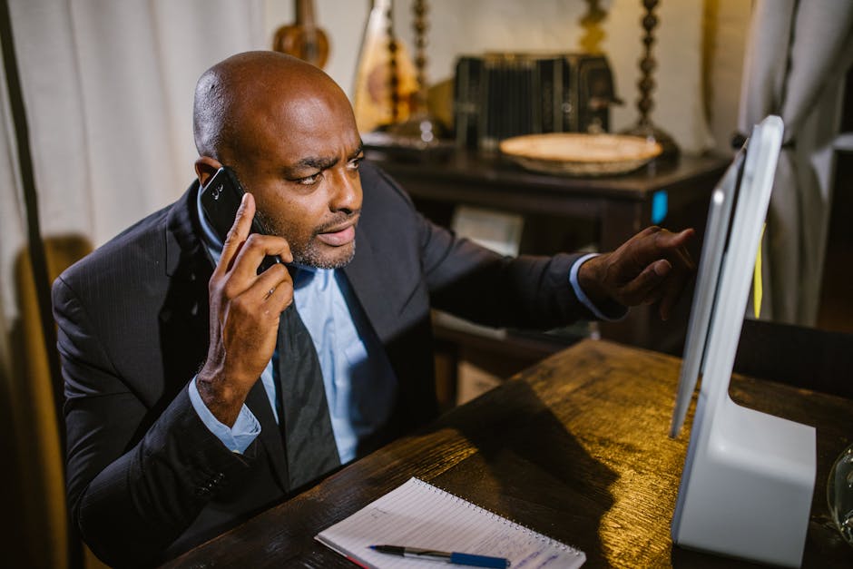 Professional man in suit talking on cellphone while working at computer indoors.