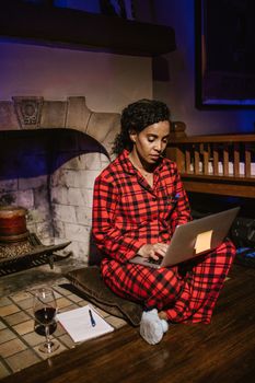 Woman in red plaid pajamas working on a laptop by a cozy fireplace with a glass of wine.