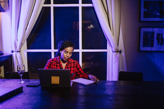 A woman working from home at night with a laptop and a glass of wine, in a cozy room setting.