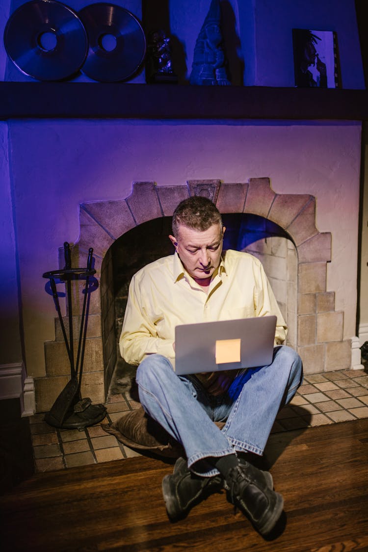 Man In Yellow Dress Shirt Sitting By A Fireplace Using Laptop