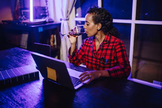 Woman in plaid shirt drinking wine while working remotely on a laptop at home.