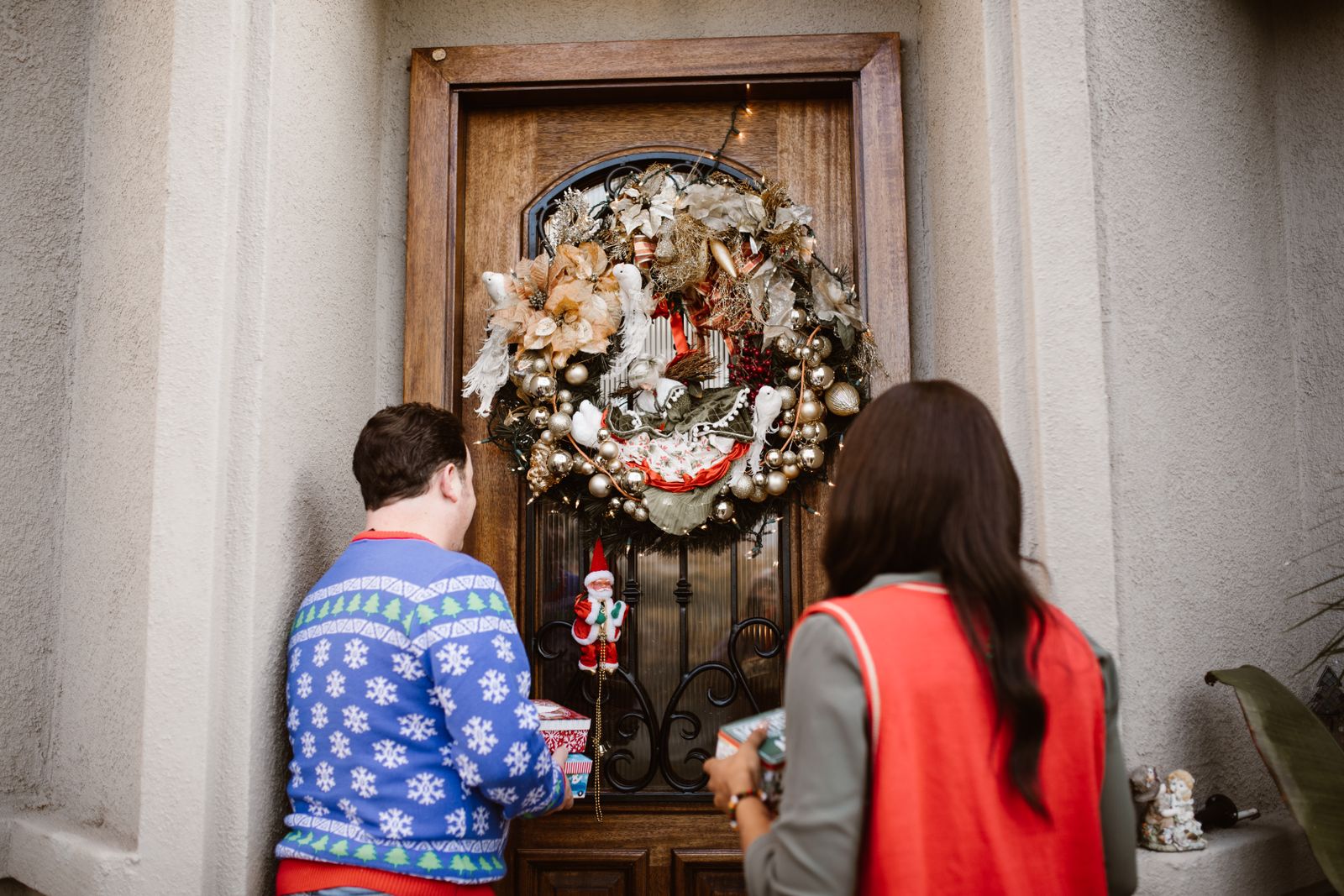 Two guests carrying gifts arriving at a front door with a large ornate Christmas wreath.