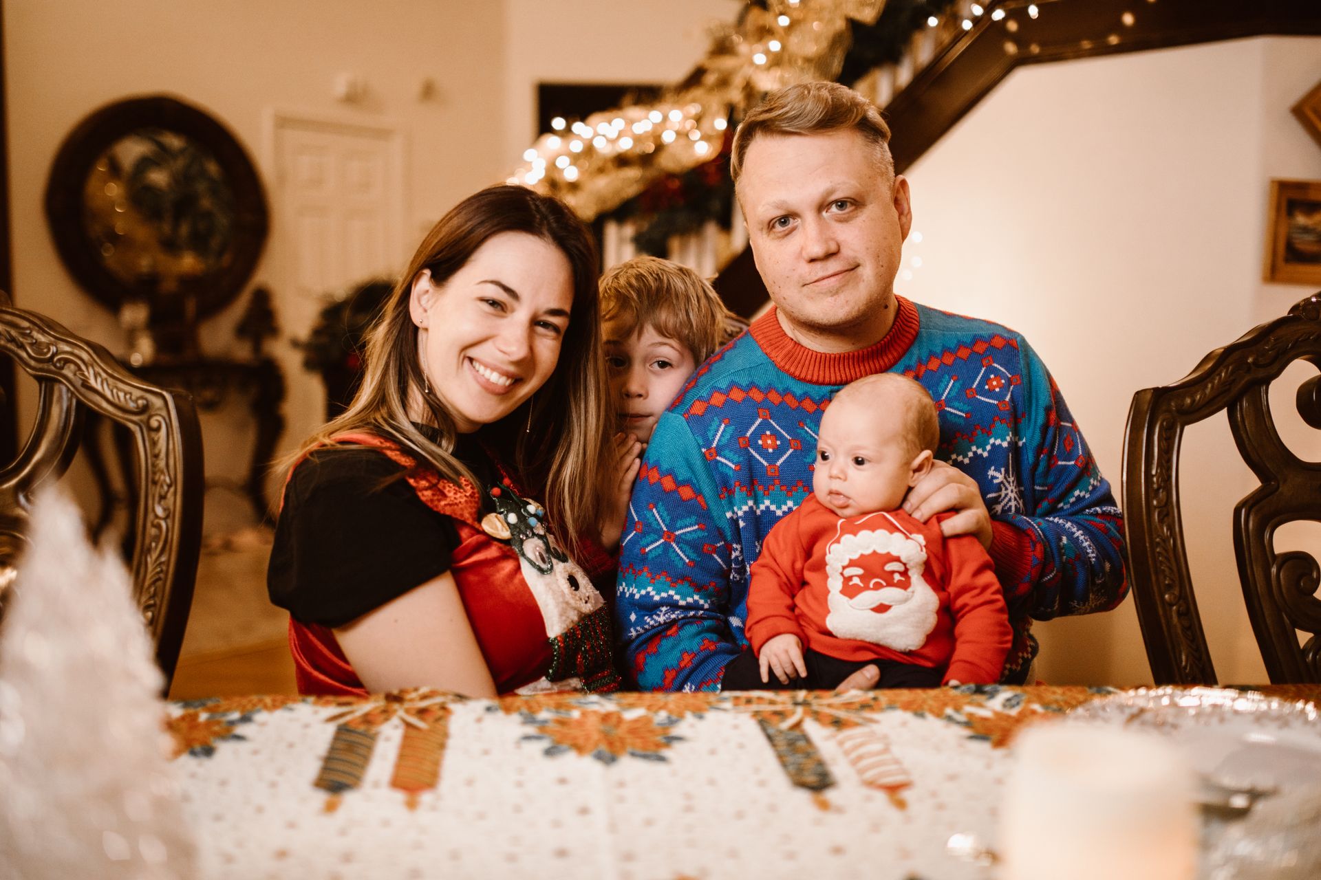 Family gathered indoors with festive Christmas sweaters, smiling around the table.