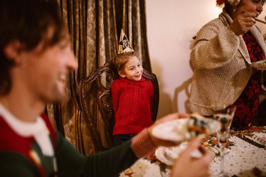 Child wearing a party hat enjoying family Christmas dinner, surrounded by happiness.