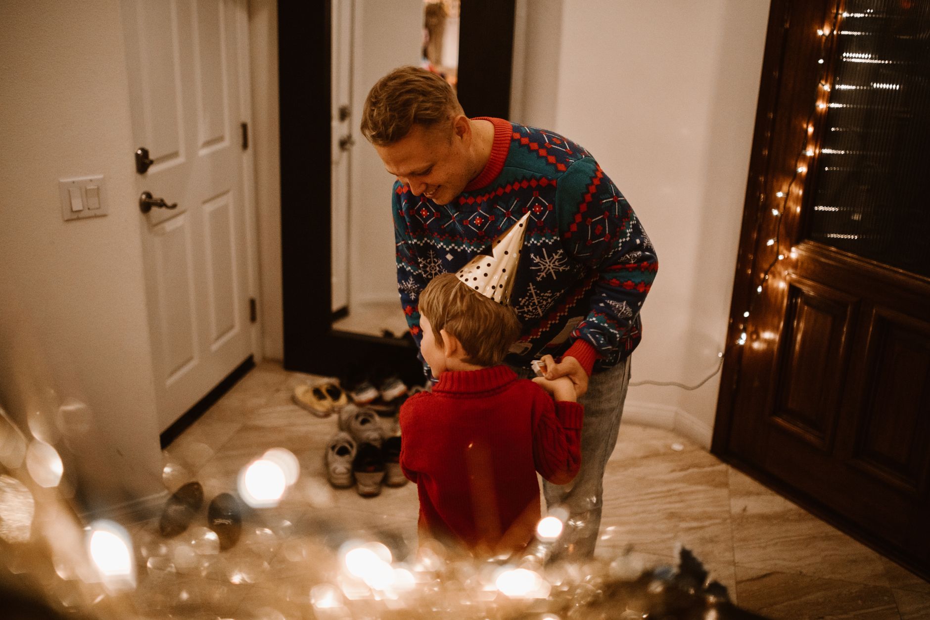 Father and son share a joyful moment in Christmas sweaters and party hats indoors.