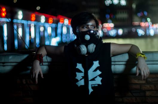 A woman in a gas mask, posing outdoors at night with city lights in the background.