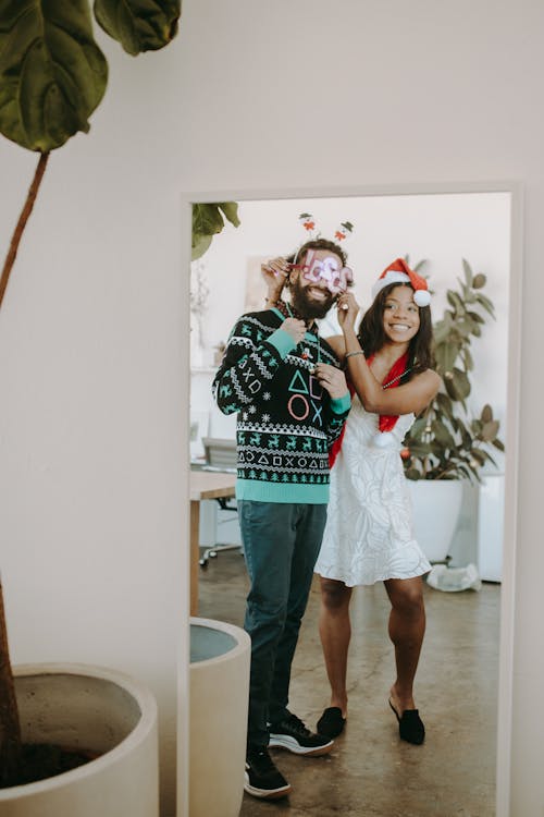 A Woman in White Dress Standing Beside a Man in Ugly Christmas Sweater while Looking at the Mirror