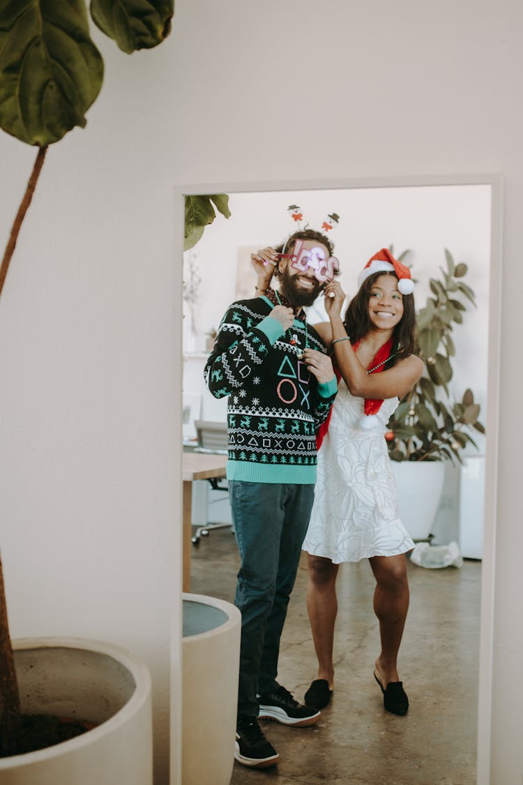 A Woman In White Dress Standing Beside A Man In Ugly Christmas Sweater While Looking At The Mirror