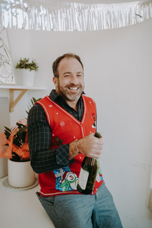 Free Smiling man in an ugly Christmas sweater holding a bottle of champagne indoors. Stock Photo