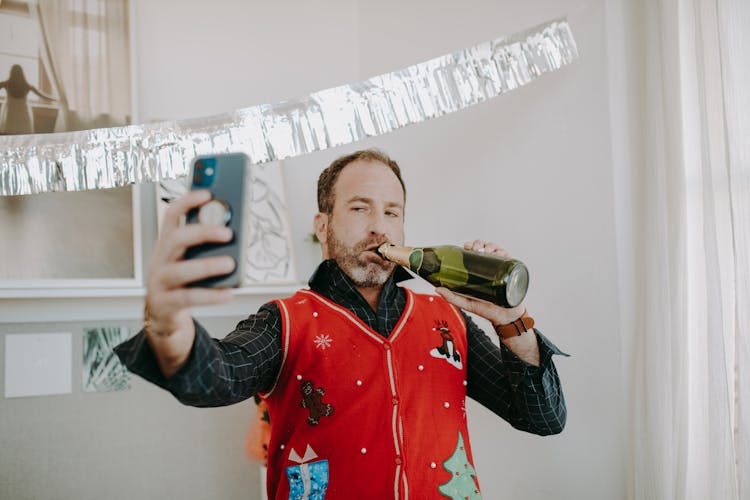 A Man In Red Ugly Christmas Sweater Holding Champagne Bottle On Lips Taking A Selfie