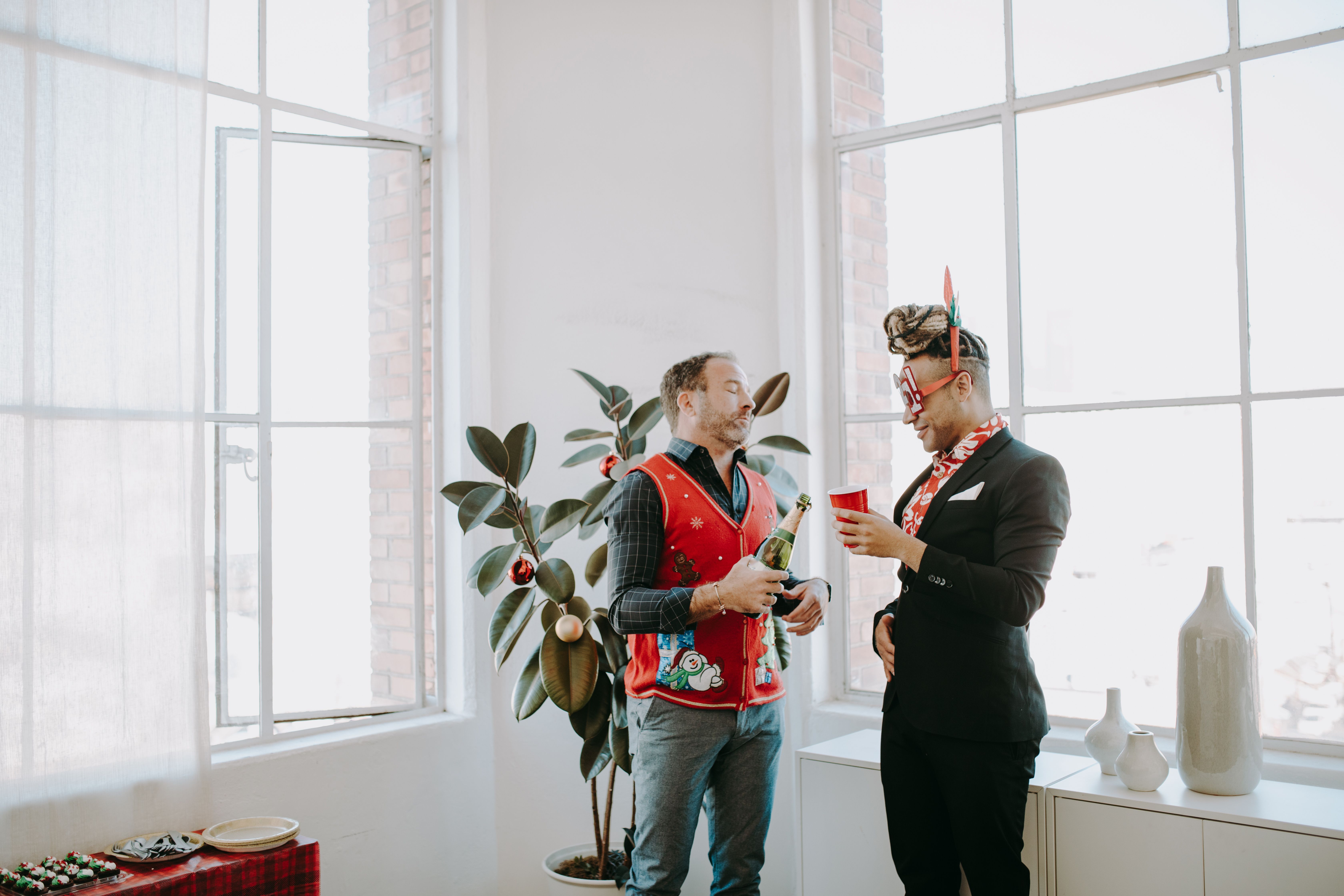 Two colleagues enjoy a holiday gathering, wearing festive ugly sweaters in a bright office setting.