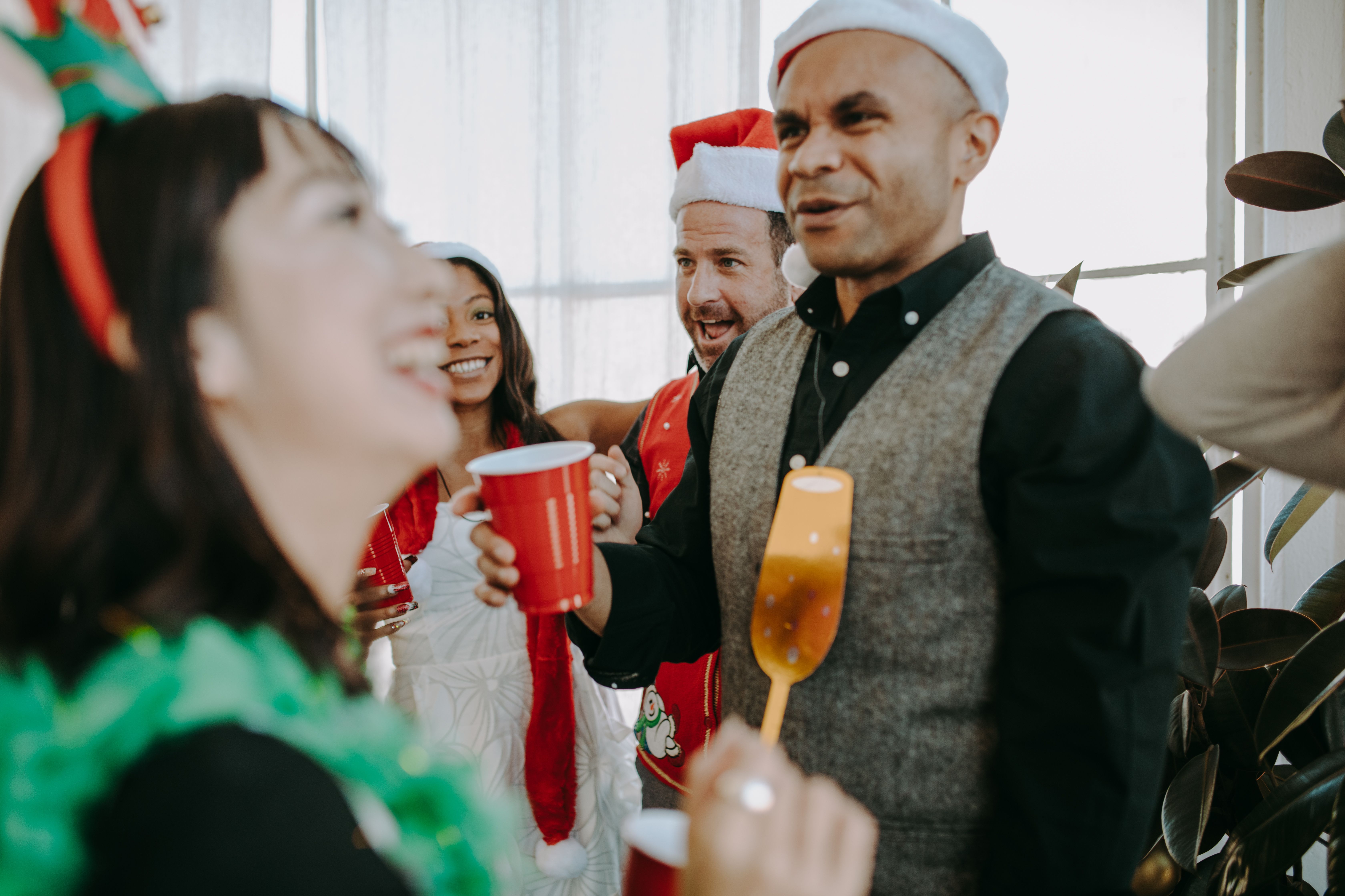 A diverse group of adults enjoying a festive holiday gathering indoors, wearing Santa hats and celebrating with red cups.