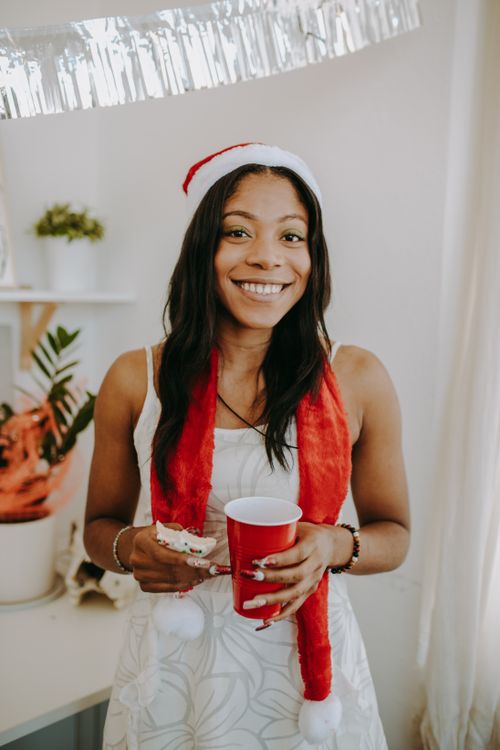 Free Smiling woman in Santa hat enjoying a holiday party indoors with a red cup and cookies. Stock Photo