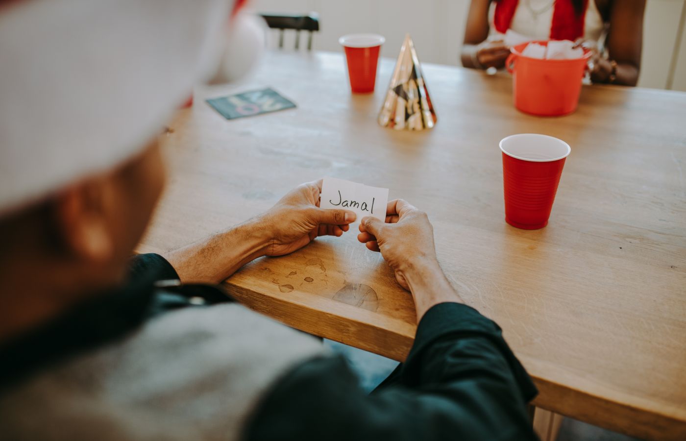 Casual indoor gathering scene with friends holding name card, red cups, and party hats on a wooden table.