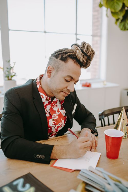 Free A young man with dreadlocks writing at a table during a festive indoor party setting. Stock Photo