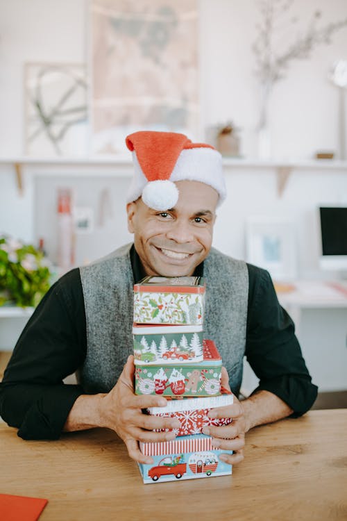 A Man Wearing Santa Hat Sitting at a Wooden Table Holding Christmas Presents 