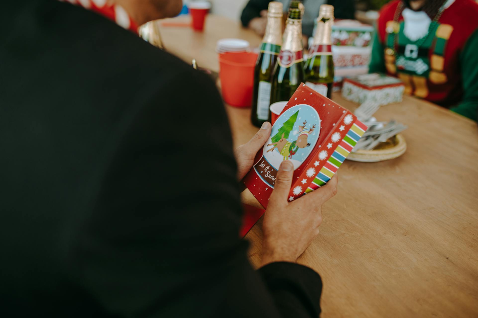 Adults holding a colorful Christmas gift during a festive holiday gathering with drinks on the table.