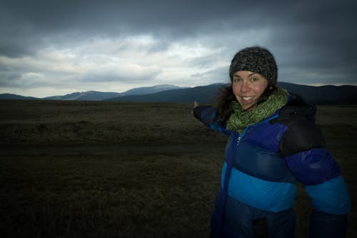 Smiling Woman Wearing Blue and Black Zip-up Bubble Jacket Pointing on Green Grass Field Under Gray Clouds