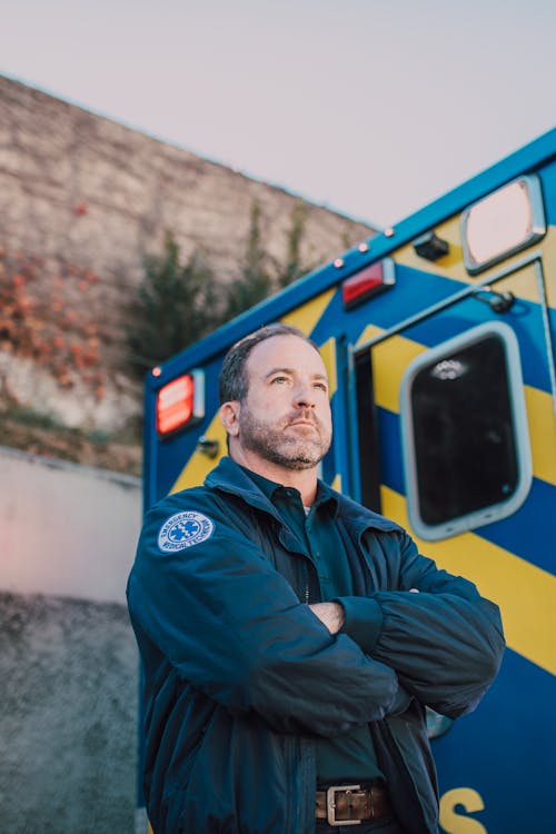 Man in Blue Jacket with Arms Crossed Standing Behind an Ambulance